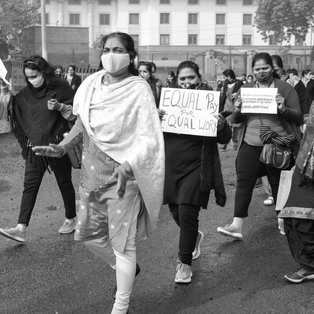 A black and white image of a women's rally advocating for equal pay, with participants wearing face masks and holding placards.