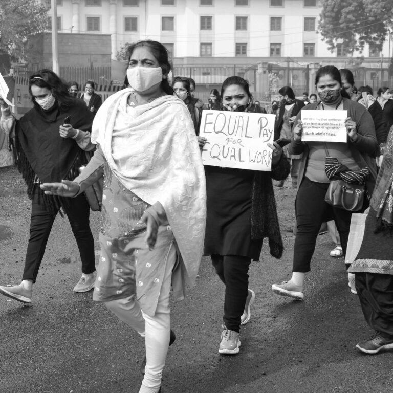 A black and white image of a women's rally advocating for equal pay, with participants wearing face masks and holding placards.